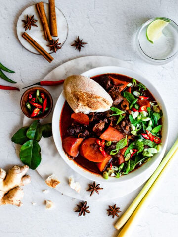 Bowlful of Bo Kho (Vietnamese Beef Stew) garnished with green onions, mint Thai basil, Thai chilies, and a baguette. Herbs and spices in background.