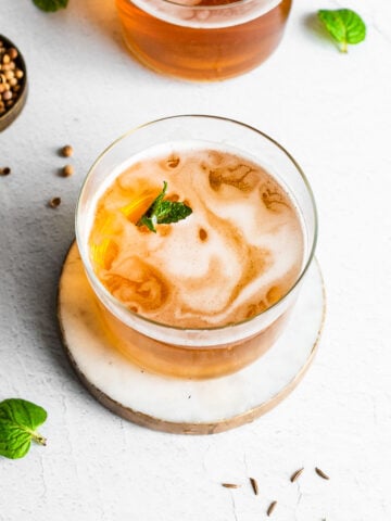 A glass of bread kvass garnished with fresh mint, fresh mint, coriander seeds and caraway seeds in background