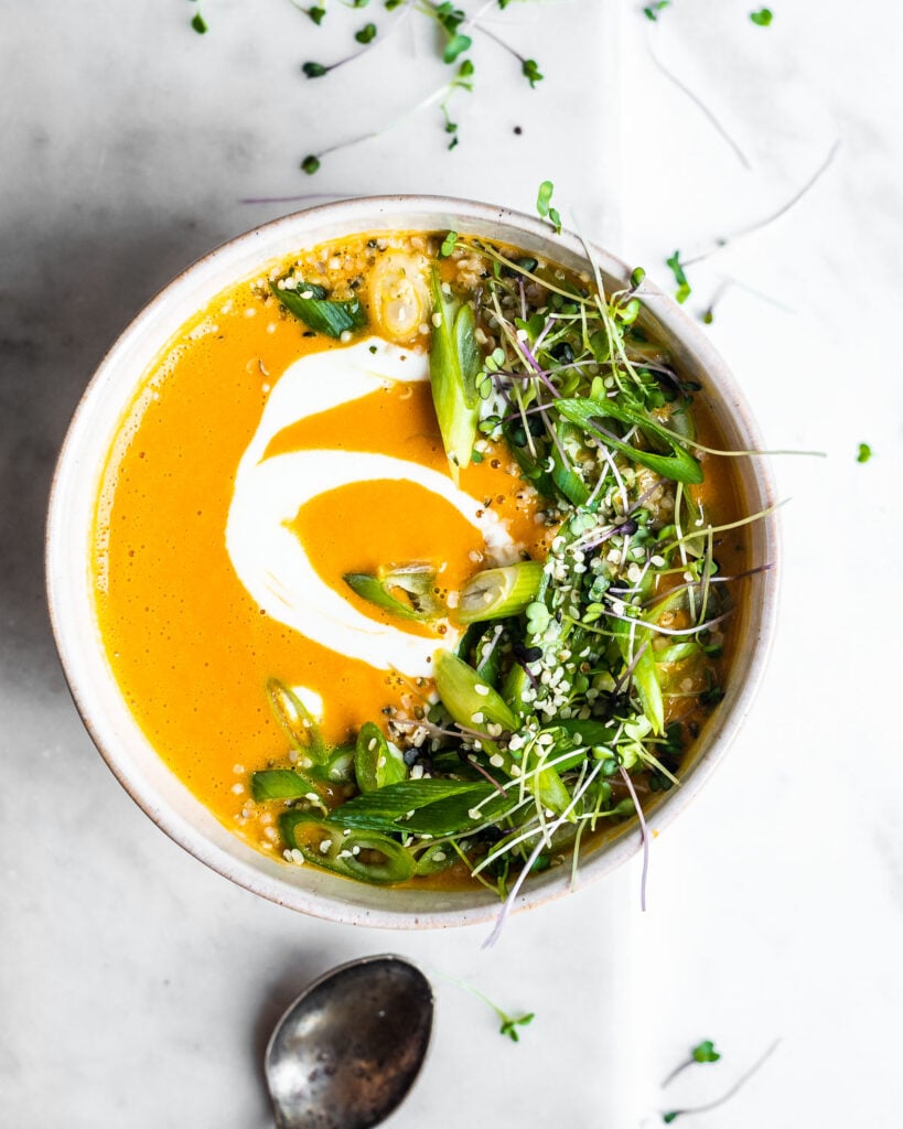 Red Kuri squash soup in a stoneware bowl, garnished with yogurt, sliced green onions, microgreens and hemp seeds.