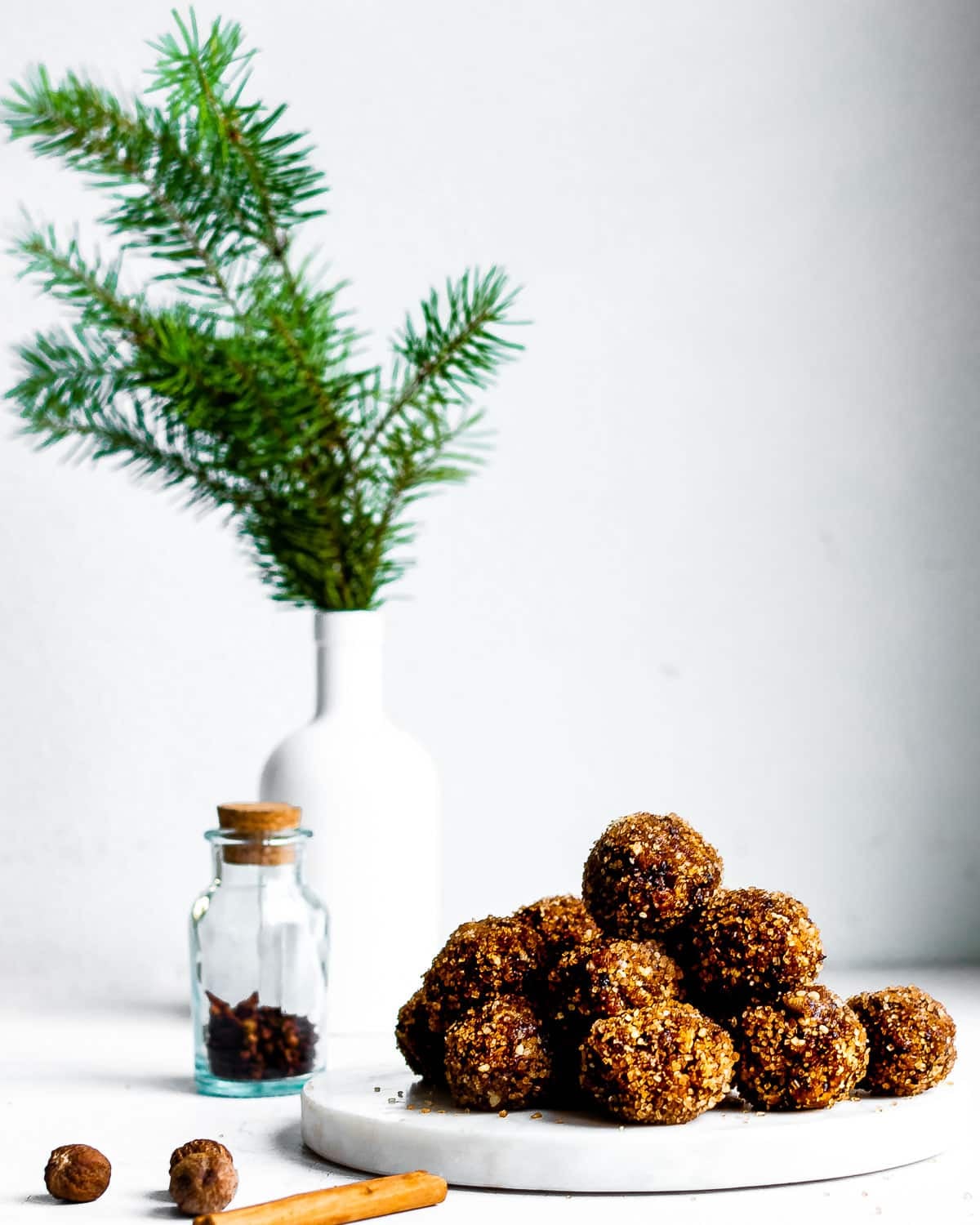 Sugar plums in a stack on a white plate, spices in foreground, fir branch in background.